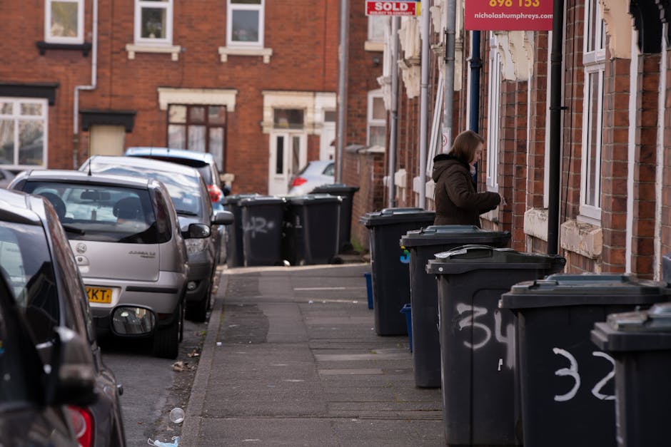 A woman dressed in a dark brown coat stands on a residential pavement beside a row of black wheelie bins, which are lined up along an exterior brick wall of a terraced house. The bins are made of plas